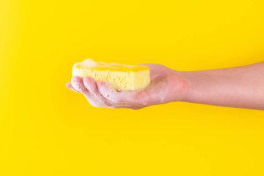 Persons hand holding yellow sponge for dish wash. Washcloth covered in soap. Domestic chores and supplies concept. Sensitive dishwashing detergent. Copy space in left side. Isolated on yellow