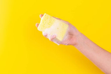 Persons hand holding yellow sponge for dish wash. Washcloth covered in soap. Domestic chores and supplies concept. Sensitive dishwashing detergent. Copy space in left side. Isolated on yellow