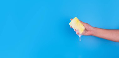 Persons hand holding yellow sponge for dish wash. Washcloth covered in soap. Domestic chores and supplies concept. Sensitive dishwashing detergent. Copy space in left side. Isolated on blue