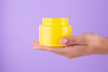 Advertising yellow container, jar in female hand on purple background. Skincare, bodycare beauty product in plastic pot package concept with vitamin c. Yellow unbranded, balsam, face or hand creme jar