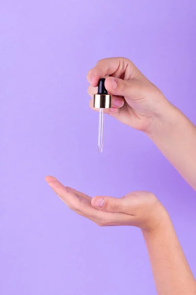 Hands of white woman holding cosmetic serum pipette on the purple background. Liquid emulsion with hyaluronic acid. Beauty cosmetic spa product
