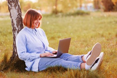 Photo of attractive lady student sit ground lean tree browsing notebook chatting friends do home tasks inspiration nature beauty harmony wear t-shirt jeans park outdoors