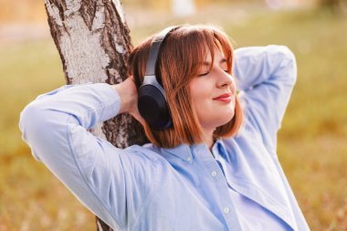 Young smiling woman listening music with headphones at sunset. Caucasian girl listening songs via wireless black headphones. Closeup face of teen and keeps the rhythm of the song.