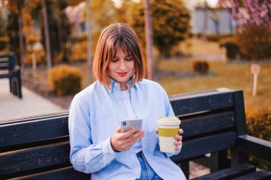Photo of charming pretty young lady communicating modern gadget 5G with cup of tea coffee sitting bench smiling outside urban city stree