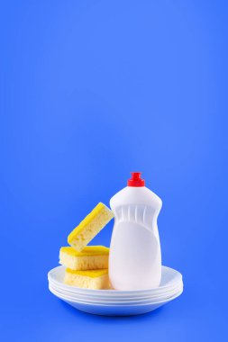 Cleaning yellow sponge, white plate and detergent for dishes on a blue background. Top view, close up. Concept cleaning company, purity, cleaning, kitche