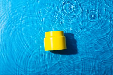 The yellow jar of moisturizing cream in the blue water wave and water drops on the blue water background. Blank label for branding mock-up. Summer water pool fresh concept. Flat lay, top view