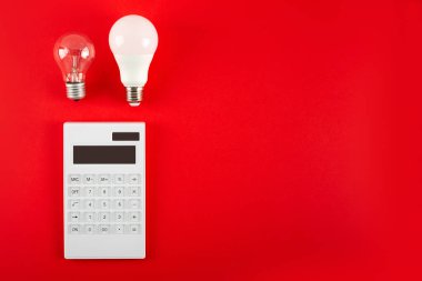 Beyaz hesap makinesi ve parlak lamba ya da kırmızı arka planda LED ampul. Elektrik faturalarının ödenmesi kavramı. Tasarruf elektriği kavramı. Faturaların ödenmesinde indirim