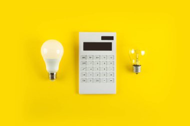Beyaz hesap makinesi ve parlak lamba ya da sarı arka planda LED ampul. Elektrik faturalarının ödenmesi kavramı. Tasarruf elektriği kavramı. Faturaların ödenmesinde indirim