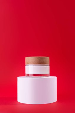 Beyaz, cam krema kavanozu tezgahta, beyaz podyum kırmızı arka planda. İlanlar, el ilanları, afişler veya dergi geçmişi için Moda Kozmetik Ürünlerinin Tasarım Şablonu. 