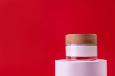 Beyaz, cam krema kavanozu tezgahta, beyaz podyum kırmızı arka planda. İlanlar, el ilanları, afişler veya dergi geçmişi için Moda Kozmetik Ürünlerinin Tasarım Şablonu. 