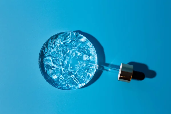 Damlalık pipetli açık başlık, daire şeklinde serum, cam podyum. Mavi arka plan, gün ışığı ve jel dokusunun görünüşü. Yüz ve vücut arabası için güzellik konsepti.