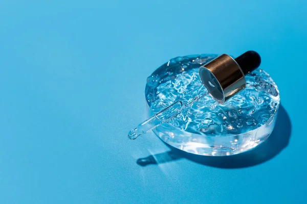 Damlalık pipetli açık başlık, daire şeklinde serum, cam podyum. Mavi arka plan, gün ışığı ve jel dokusunun görünüşü. Yüz ve vücut arabası için güzellik konsepti.