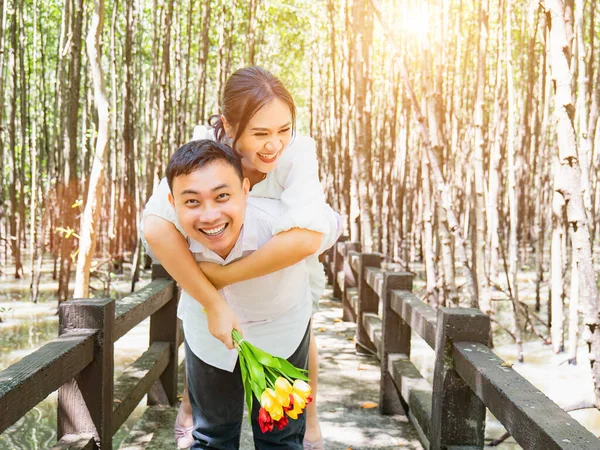 一位身穿白衣 手持一束鲜花的亚洲女人 幸福地骑在一位亚洲男子的背上 在红树林中 在被求婚后带着凉爽的阴影示爱和幸福 — 图库照片