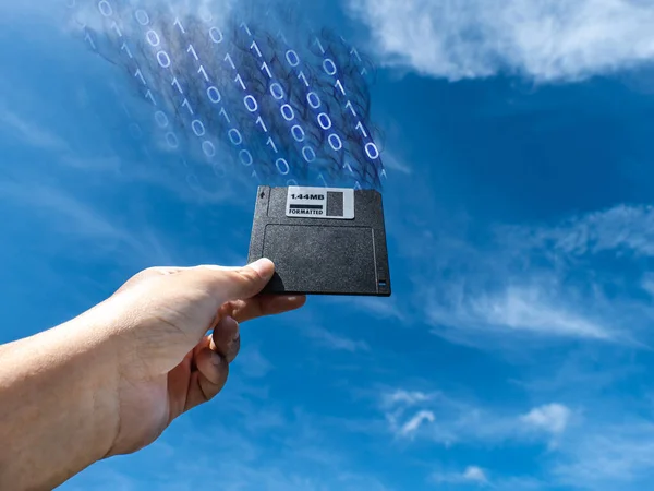 Hand holding a Floppy A disc, insert the reader. to read and write data It is an old technology with a square shape. the background is the sky There is digital information flowing out.