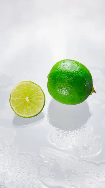 The green lime is blank with the cut lime slice showing the inside of the wet lemon pulp on a clear glass surface, reflecting the shadows of the lime and the wet water, giving it its freshness.