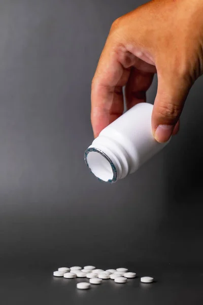 Hand holding unlabeled white plastic pill bottle open cap Poured out several small white pills. come out below in black background