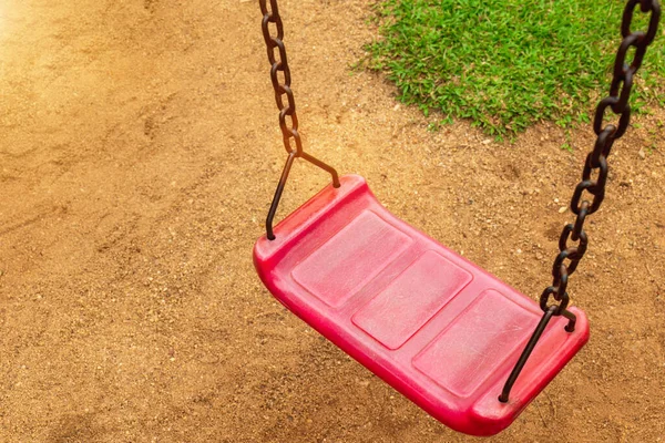 A red swing is attached to two old rusted chains in the playground. in the park saw the ground and the grass