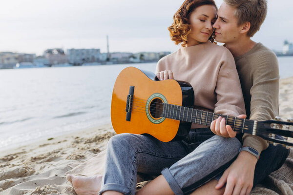 young guy teaching his girlfriend how to play guitar, self-taught. Couple in love having fun on the beach hugging and kissing