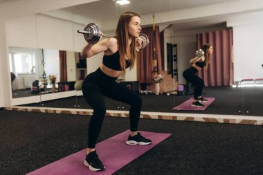 Halterle hamle yapan bir kız. Spor giyim kuşamında kaslı vücutlu, esmer bir kadın karanlık bir atmosferde spor salonunda bacaklarını çalıştırıyor. Vücut geliştirme kavramı.