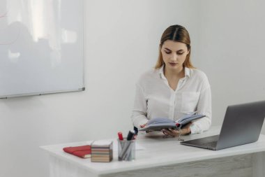 Genç öğretmen bilgisayarda çalışıyor. O, beyaz bir gömlek giyiyor. Öğrenciler için çevrimiçi eğitim veriyor. O bir defter tutuyor. Online dersler. Giyin.