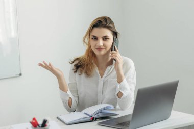 Akıllı telefondan konuşan genç bir iş kadınının portresi, bir dizüstü bilgisayarla çalışmak ve ofisteki bir deftere yazı yazmak. Kız kollarını açar Evdeki bilgisayar masasında güzel bir kız.