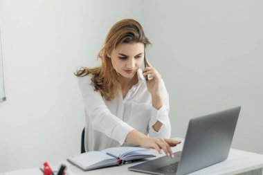 Akıllı telefondan konuşan genç bir iş kadınının portresi dizüstü bilgisayarda çalışıyor ve ofisteki deftere yazı yazıyor. Evdeki bilgisayar masasında güzel bir kız. İnternet üzerinden bir iş kadını başlat..