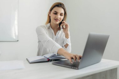 Akıllı telefondan konuşan genç bir iş kadınının portresi dizüstü bilgisayarda çalışıyor ve ofisteki deftere yazı yazıyor. Evdeki bilgisayar masasında güzel bir kız. İnternet üzerinden bir iş kadını başlat..
