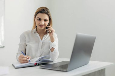 Akıllı telefondan konuşan genç bir iş kadınının portresi dizüstü bilgisayarda çalışıyor ve ofisteki deftere yazı yazıyor. Evdeki bilgisayar masasında güzel bir kız. İnternet üzerinden bir iş kadını başlat..