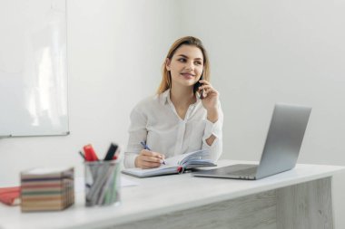 Akıllı telefondan konuşan genç bir iş kadınının portresi dizüstü bilgisayarda çalışıyor ve ofisteki deftere yazı yazıyor. Evdeki bilgisayar masasında güzel bir kız. İnternet üzerinden bir iş kadını başlat..