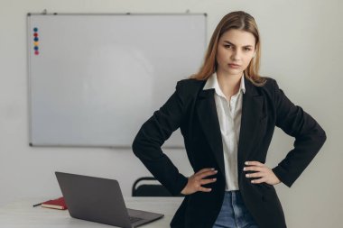 Tatmin olmamış bir öğretmenin portresi. Ellerini kemerinde tutuyor. Kız beyaz gömlek ve siyah ceket giyiyor.