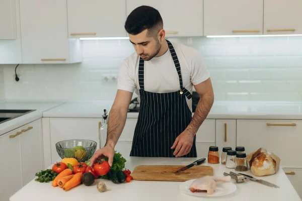 Mutfakta yakışıklı bir adamın portresi. Siyah önlük giymiş bir adam. Yemek yapmak için sebze doğra.
