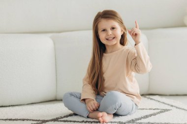 Küçük tatlı bir kızın portresi. O, kanepenin yanında yerde oturuyor. Kız kameraya bakar ve parmağını kaldırır.