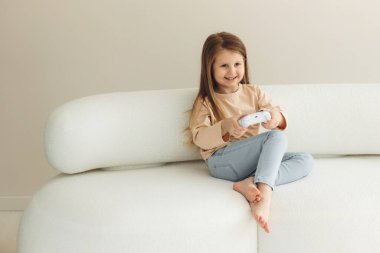 Oturma odasındaki kanepede otururken elinde oyun kumandasıyla video oyunu oynayan küçük bir kız.