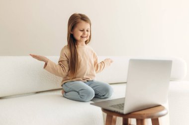 Modern teknolojiye bağımlı, elinde bilgisayarla kanepede oturan neşeli küçük tatlı kız, online oyunlardan zevk alıyor, sosyal ağlarda sosyalleşiyor, komik video içerikleri izliyor.