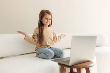 Modern teknolojiye bağımlı, elinde bilgisayarla kanepede oturan neşeli küçük tatlı kız, online oyunlardan zevk alıyor, sosyal ağlarda sosyalleşiyor, komik video içerikleri izliyor.