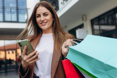 Alışveriş çantalarını tutan ve alışveriş merkezinden çıkarken akıllı telefon kullanan zarif genç bir kadının portresi.