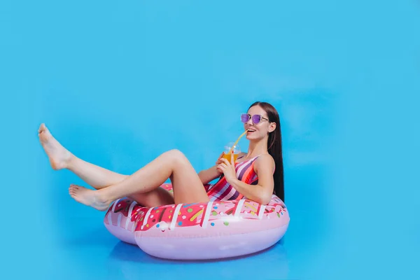 Stunning young brunette girl in colored one-piece swimsuit with sunglasses. People summer vacation vacation lifestyle concept. Sit in a swimming inflatable ring to hold with a cocktail. Photo on a blue background