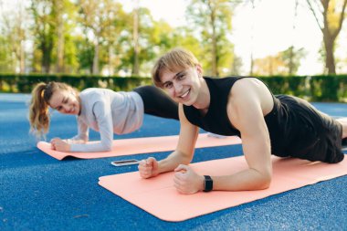 Genç fitness kadını ve erkek birlikte bir stadyumda tahta egzersizleri yapıyorlar.