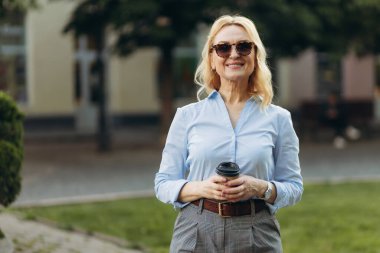 Portrait of a senior business woman holding a paper cup. Elderly smiling happy woman in her 50s walks in the city. Business Concept Job Success