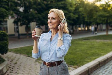 Portrait of senior business woman hold the paper cup and talking phone in the city. Happy mature business woman 50s