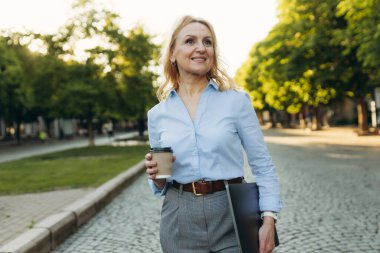 Portrait of a successful mature businesswoman wearing stylish clothes. Woman in her 50s. Woman holding laptop and paper cup of coffee while walking in the city. Lifestyle and work concept