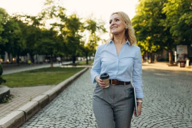 Portrait of a successful mature businesswoman wearing stylish clothes. Woman in her 50s. Woman holding laptop and paper cup of coffee while walking in the city. Lifestyle and work concept