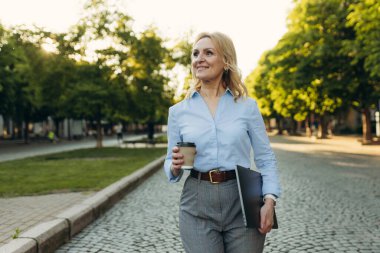 Portrait of a successful mature businesswoman wearing stylish clothes. Woman in her 50s. Woman holding laptop and paper cup of coffee while walking in the city. Lifestyle and work concept