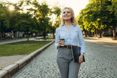 Portrait of a successful mature businesswoman wearing stylish clothes. Woman in her 50s. Woman holding laptop and paper cup of coffee while walking in the city. Lifestyle and work concept