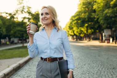 Portrait of a mature woman in stylish clothes. Women of the 50s. Woman holding laptop and drinking coffee while walking around the city. Lifestyle and work concept