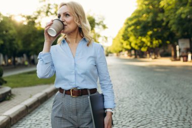 Portrait of a mature woman in stylish clothes. Women of the 50s. Woman holding laptop and drinking coffee while walking around the city. Lifestyle and work concept