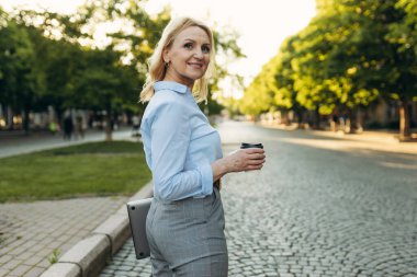 Portrait of a successful mature businesswoman wearing stylish clothes. Woman in her 50s. Woman holding laptop and paper cup of coffee while walking in the city. Lifestyle and work concept