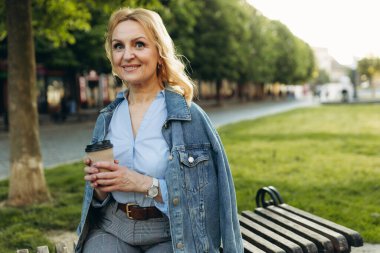 Happy mature woman wearing stylish clothes. Woman 50 years old. Woman resting on a bench and drinking coffee in the city. Lifestyle concept