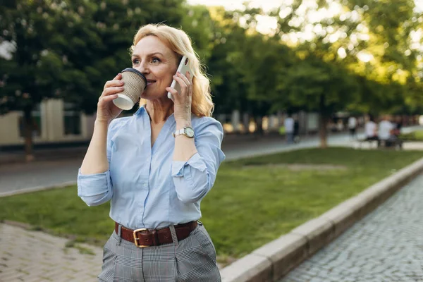 stock image Portrait of senior business woman hold the paper cup and talking phone in the city. Happy mature business woman 50s