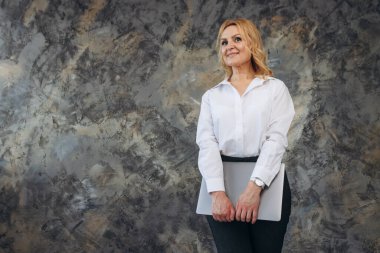 Elinde dizüstü bilgisayarla gülümseyen olgun bir kadının portresi.